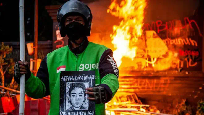 一名年轻人戴黑色口罩、佩戴头盔站在焚烧的建筑物前，手里拿着一个肖像画。
